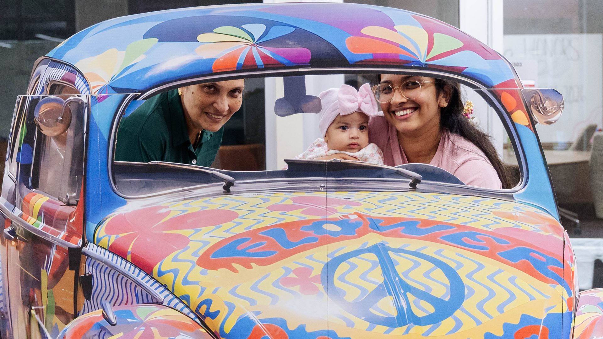A family with an infant smiles at the camera from inside a Volkswagen Beatle decorated in 1960s style graphics and a peace sign